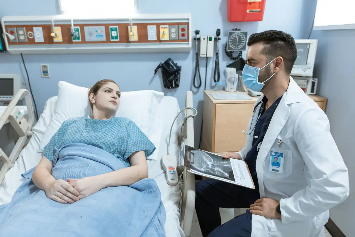 Medic cu mască discută cu o pacientă întinsă în patul din salon
