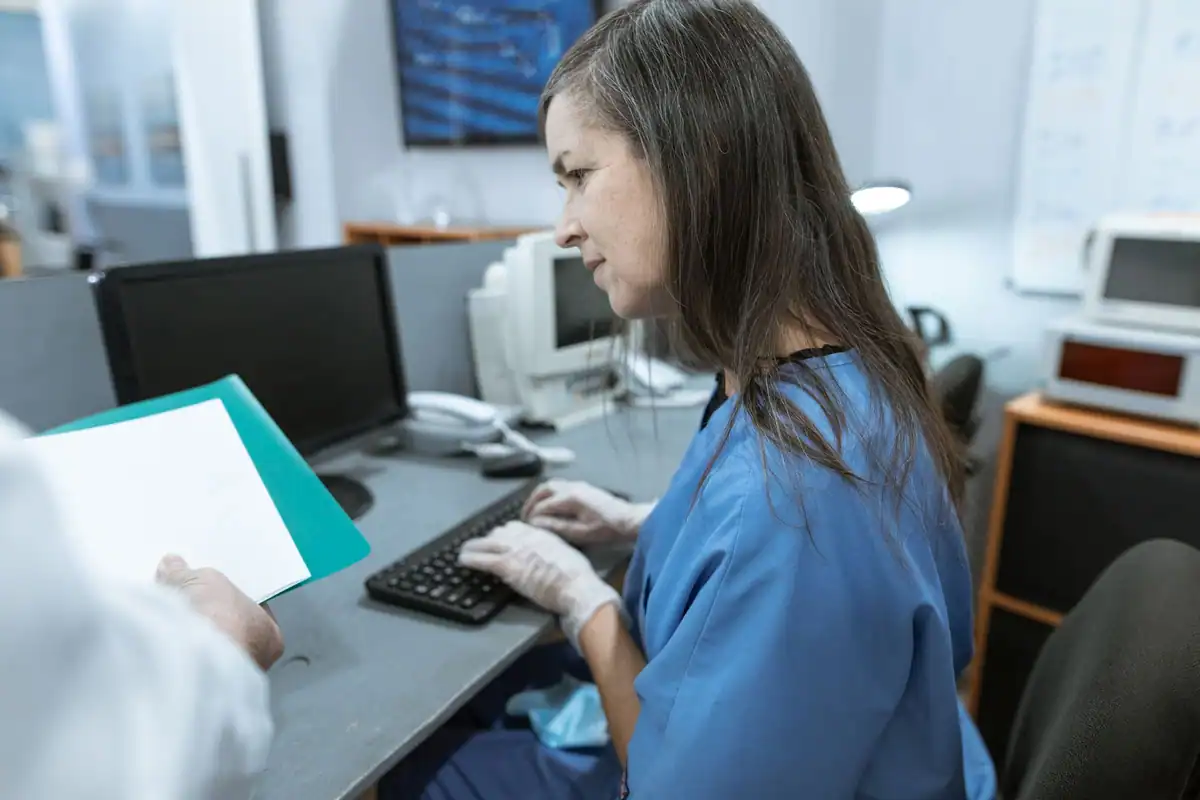 Asistentă tastează la calculator în cabinet, în timp ce cineva îi dă un document