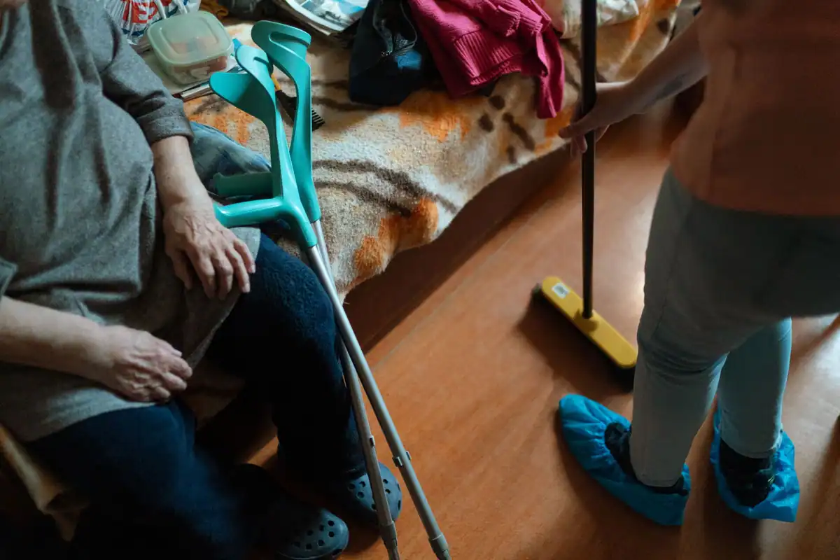 Persoană cu cârje stă pe scaun, în timp ce lângă ea se face curățenie