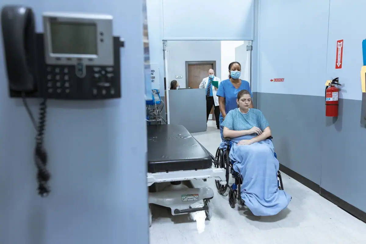 Pacientă în scaun cu rotile este condusă pe holul spitalului, lângă un pat