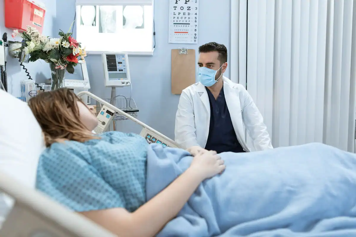 Pacientă în pat de spital stă de vorbă cu un medic cu mască
